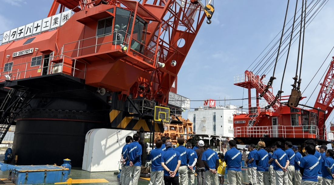 建設現場で赤いクレーンの前に青い制服を着た作業員が集まっている様子。