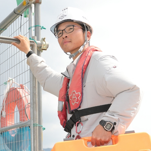 建設現場でヘルメットとライフジャケットを着用した男性作業員がツールボックスを持っている写真。