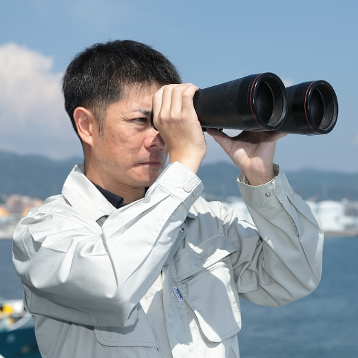 船の上で双眼鏡を覗く作業服の男性の写真