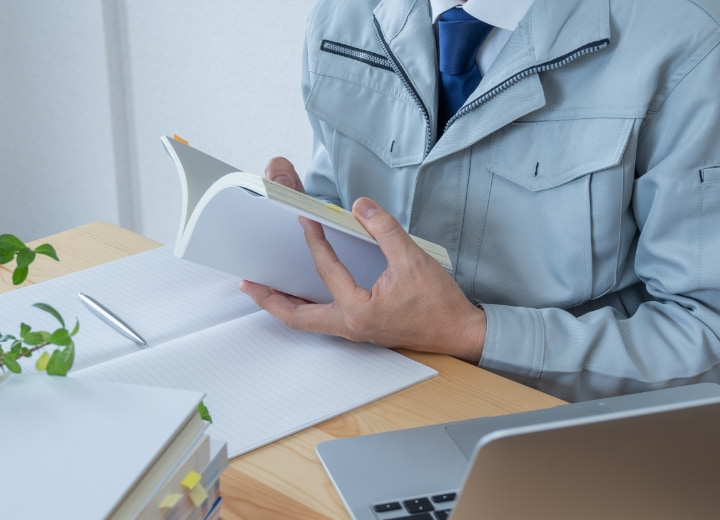 作業服を着た男性が本を読みながら仕事をしているデスクの様子。