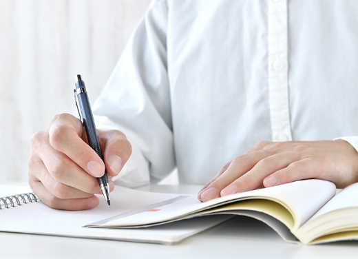 ノートにペンで書き込む白いシャツの人物の手元の写真。