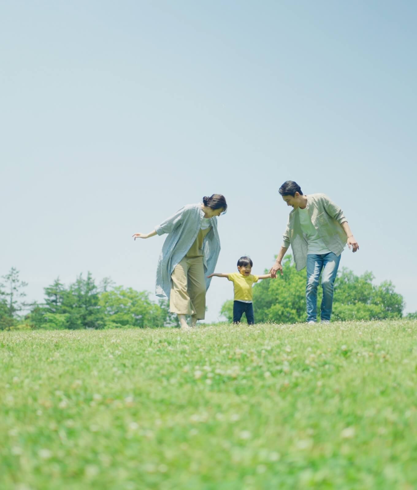 家族が子供と一緒に野原を歩いている様子。青空の下、楽しそうに笑顔を交わしている。